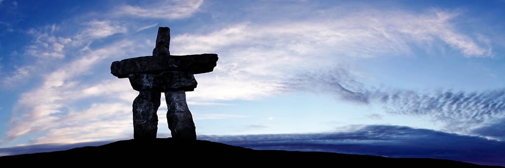 inuksuk in the foreground on a hill in the shadows with the sky and clouds in the background