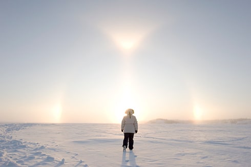 We see a person walking away from the camera dressed in a white winter jacket and black winter pants. They're walking in an open area by themselves on a sunny winter day.